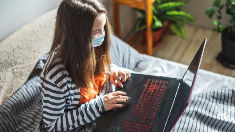 Młoda dziewczyna w masce ochronnej siedzi na łóżku w domu i używa laptopa