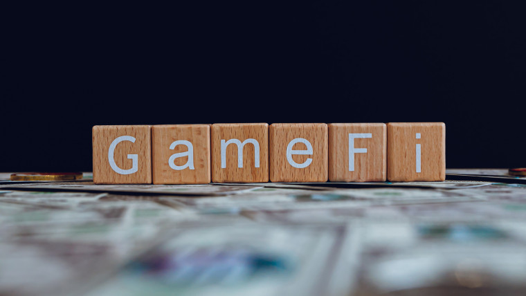 Drewniane klocki z tekstem GameFi na czarnym tle i banknotami kryptograficznymi rozrzuconymi na gro