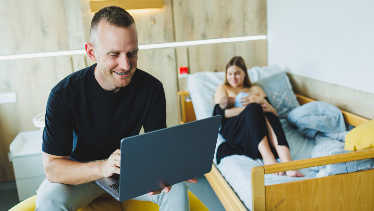 Młody mężczyzna pracuje na laptopie na tle swojej żony, która karmi swoje nowo narodzone dziecko Piersią Praca online na laptopie