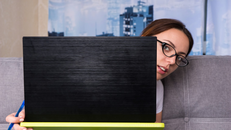 Młoda kobieta w okularach zerkająca zza laptopa, pracująca na laptopie, prowadząca interesy z domu, trzymająca długopis, gdy czyta informacje na ekranie