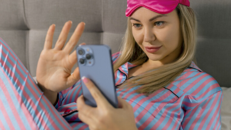 Portret pięknej radosnej kobiety w piżamie za pomocą telefonu komórkowego i uśmiechniętej leżącej na łóżku po śnie lub drzemce