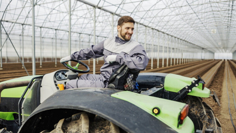Uśmiechnięty rolnik prowadzący ciągnik z pługiem i przygotowujący glebę do sadzenia w szklarni