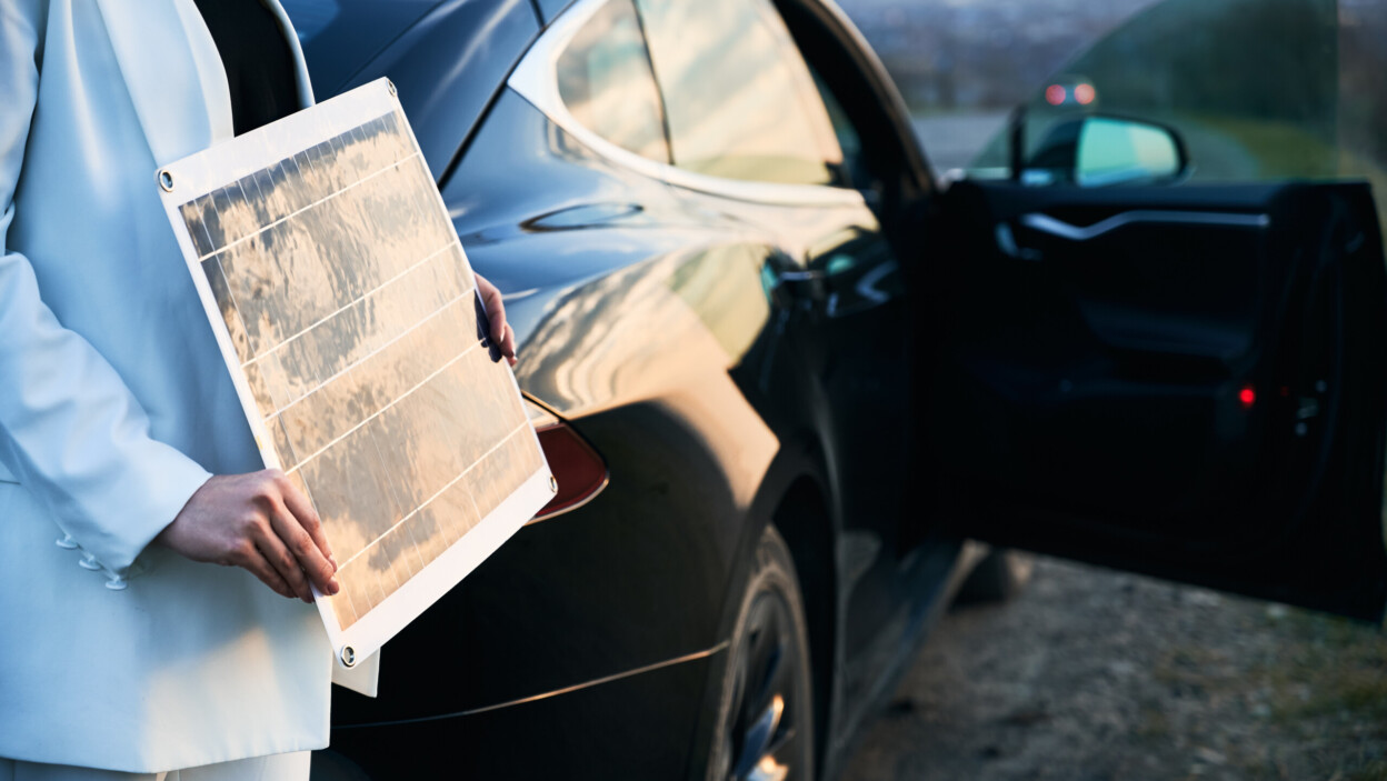 Młoda kobieta stojąca w pobliżu samochodu elektrycznego i trzymająca panel słoneczny
