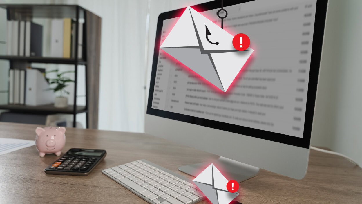 Phishing – monitor komputera wyświetla alert e-mail z ikonami ostrzegawczymi, otoczony klawiaturą, kalkulatorem, skarbonką i biurkiem w nowoczesnym biurze