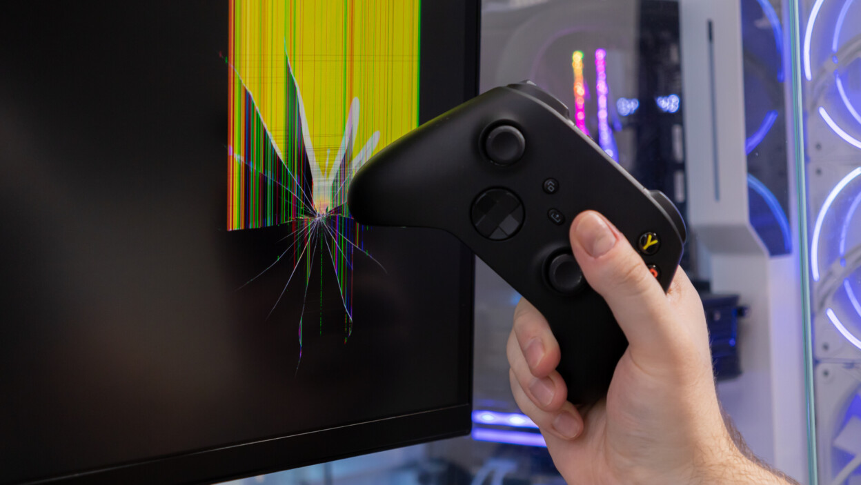 A gamer39s hand holds a black controller near a severely broken computer monitor depicting intense gaming frustration and damaged technology