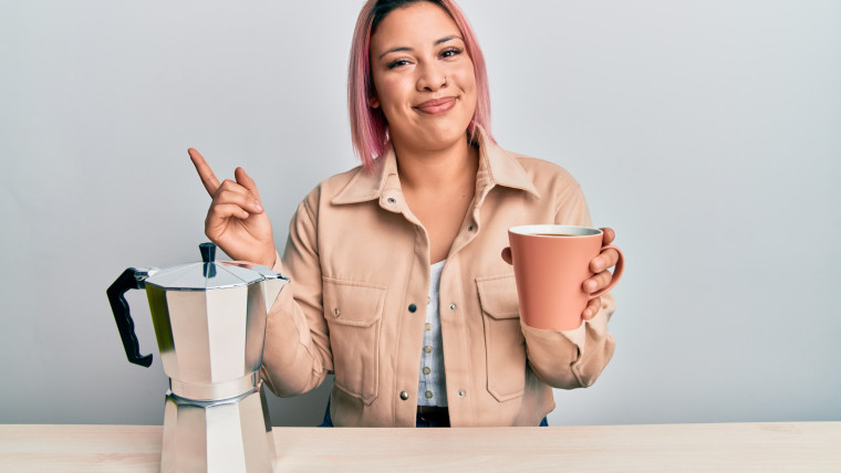 Latynoska kobieta z różowymi włosami pije filiżankę włoskiej kawy uśmiechając się radośnie, wskazując ręką i palcem na bok