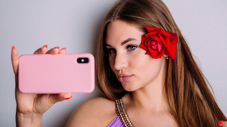 Atrakcyjna młoda kobieta robi selfie na swoim smartfonie, wydymając wargi i prostując włosy czerwonym kwiatem róży.